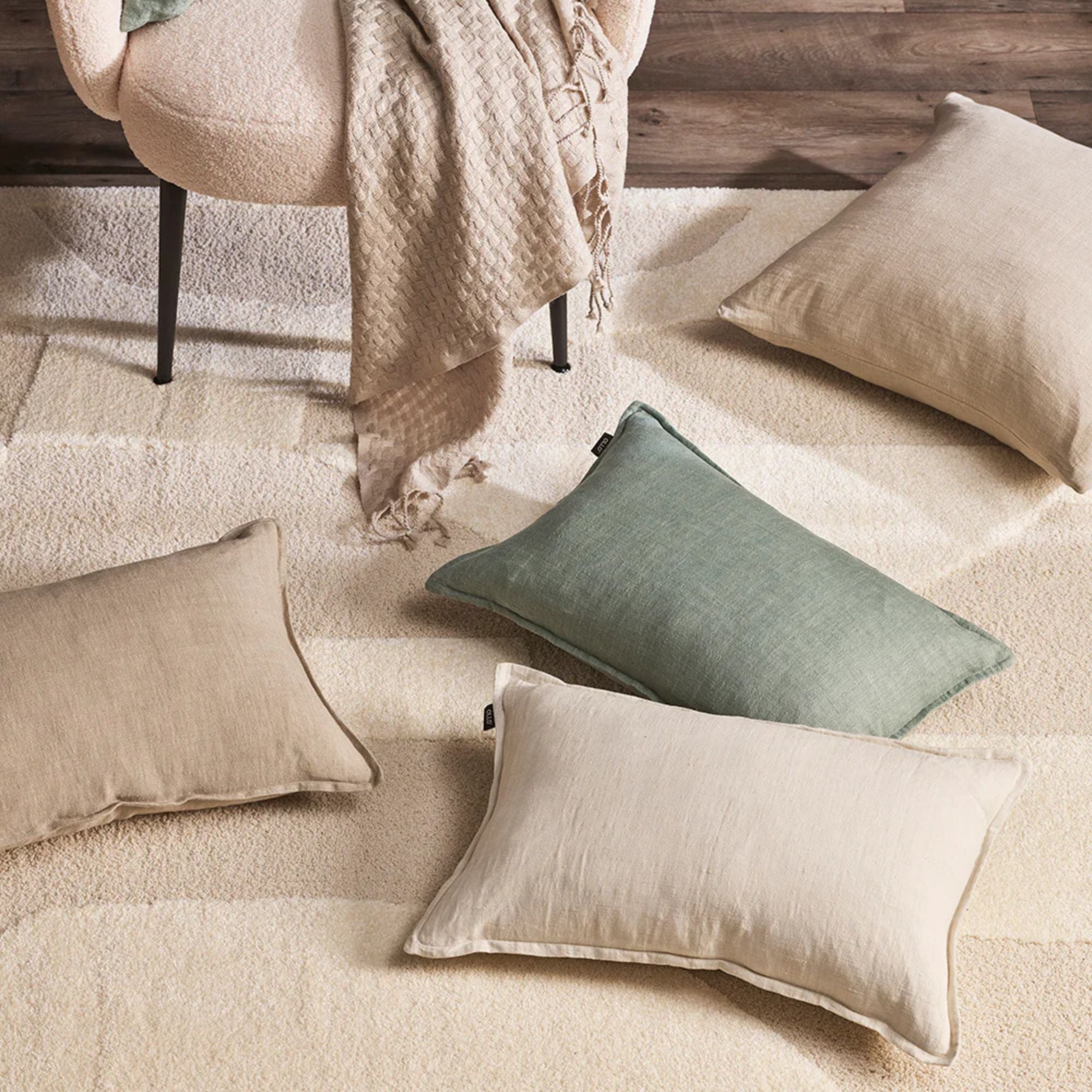 Decorative pillows in beige, green, and white on a textured surface with a chair and blanket in the background.