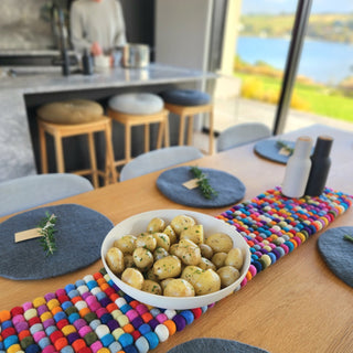 Colourful felt ball table runner styled on a wooden dining table with grey placemats and a bowl of potatoes