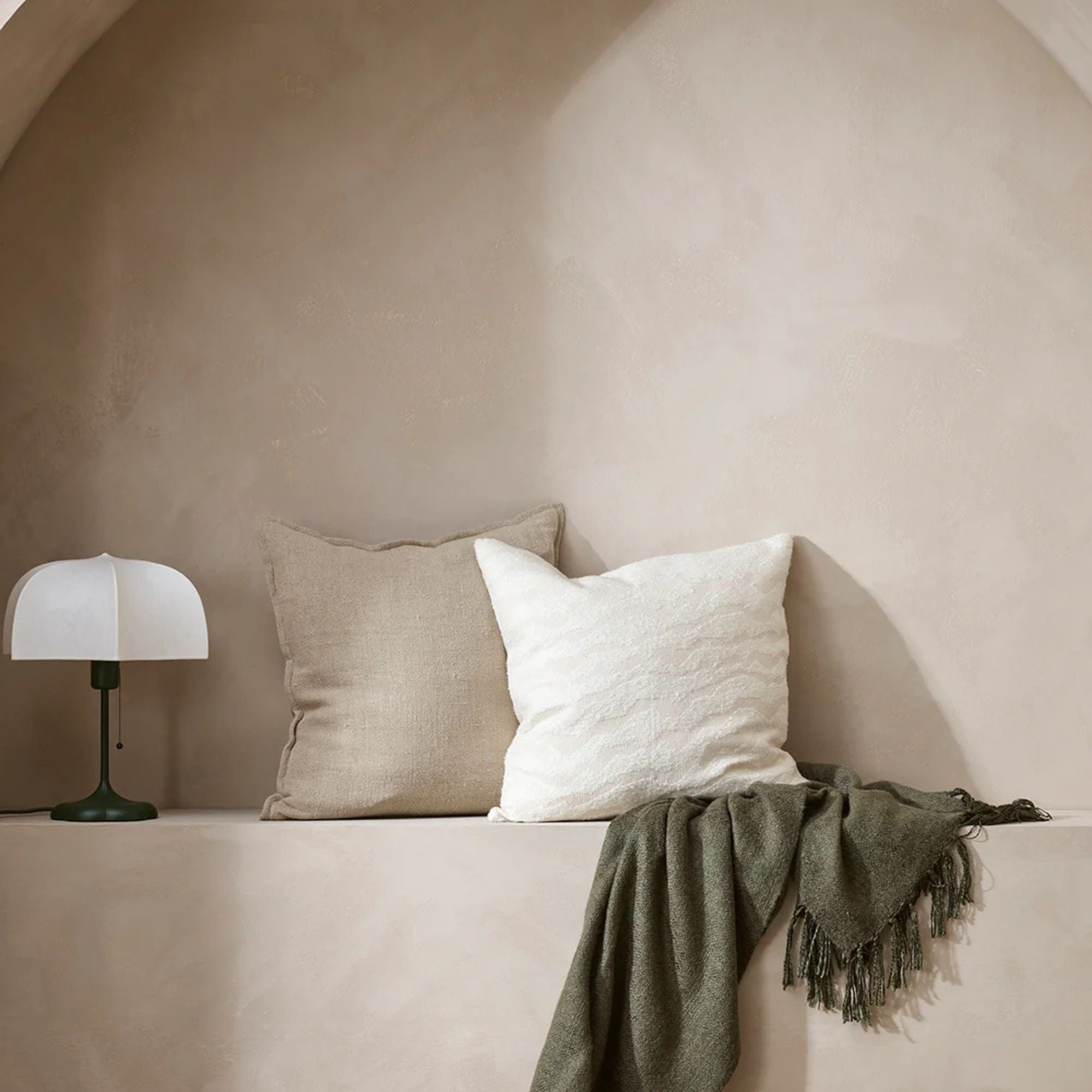 Two pillows on a ledge with a lamp and green blanket against a beige wall.