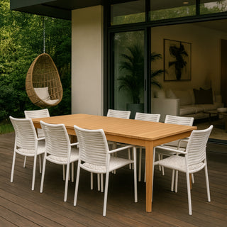Wooden outdoor dining table with white chairs on a deck, surrounded by greenery.