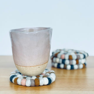Ceramic cup resting on a neutral felt ball coaster with a matching stack of coasters in the background on a wooden table.