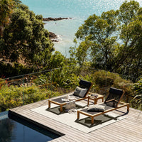 Two lounge chairs on a wooden deck overlooking a scenic ocean view with lush greenery.