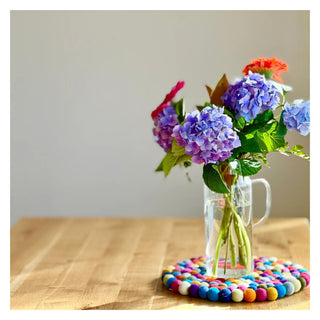 Vase of fresh flowers placed on a multicolour 30cm felt ball pot stand on a wooden table.