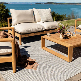 Outdoor patio set with wooden furniture and cushions by a scenic waterfront.