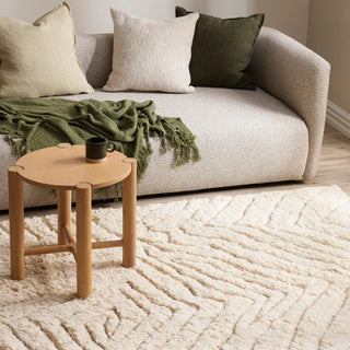 Beige sofa with cushions and a green blanket, accompanied by a wooden side table with a mug on a textured rug.