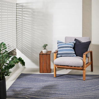 Wooden armchair with cushions on a rug in a room with white walls and a plant.