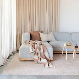 Light gray sofa with a plaid blanket in a living room setting with beige curtains and rug.