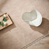 White ceramic pitcher on a beige rug with a wooden tray and flowers.