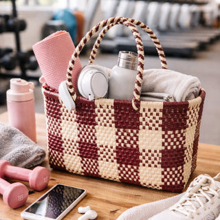 Woven basket with gym items including a water bottle, towel, and dumbbells on a wooden surface.