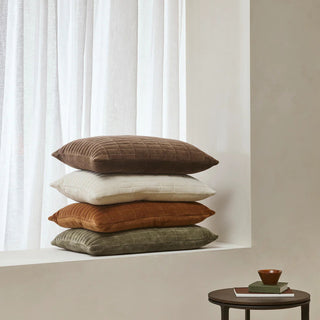 Stack of four textured pillows in brown, white, orange, and green against a neutral wall.