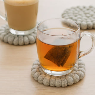 Two glass mugs with coffee and tea resting on round grey felt ball coasters on a wooden table.