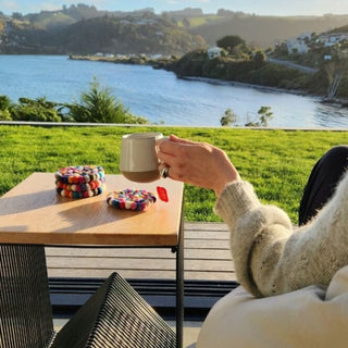 Person enjoying a hot drink outdoors by the water, with colourful felt ball coasters on a wooden outdoor table.