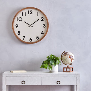 Wall clock on a light gray wall above a white console table with a plant and globe.