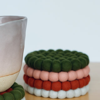 Ceramic cup beside a stack of colourful felt ball coasters in green, blush, rust, and grey on a wooden surface.